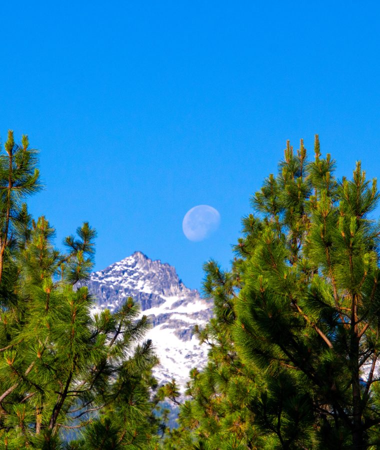 Pale moon in blue sky above rocky mountain tops.